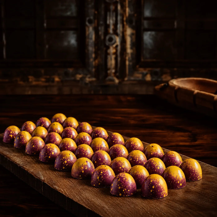 A row of glossy, dome-shaped chocolates passion fruit bon bon or confections with a purple and yellow marbled pattern and small yellow speckles, arranged on a rustic wooden board in a dark, atmospheric setting.