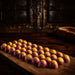 A row of glossy, dome-shaped chocolates passion fruit bon bon or confections with a purple and yellow marbled pattern and small yellow speckles, arranged on a rustic wooden board in a dark, atmospheric setting.