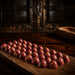 A row of glossy Raspberry chocolate truffles bon bons with a deep burgundy-red coating and yellow-orange speckles, arranged on a dark wooden board in a rustic setting.