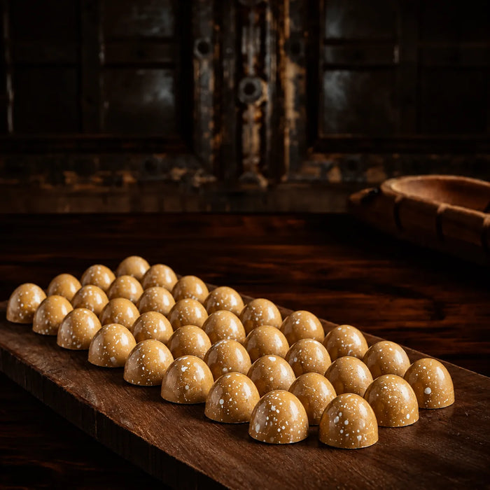 A row of glossy, dome-shaped golden-brown salted caramel chocolates with small white speckles, arranged on a dark wooden serving board in a rustic setting.