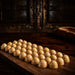 A row of glossy, dome-shaped golden-brown salted caramel chocolates with small white speckles, arranged on a dark wooden serving board in a rustic setting.