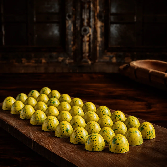 A row of small, glossy yellow confectionery Chocolate bon bons yuzu flavor with green speckles arranged on a dark wooden board in a rustic, dimly lit setting.